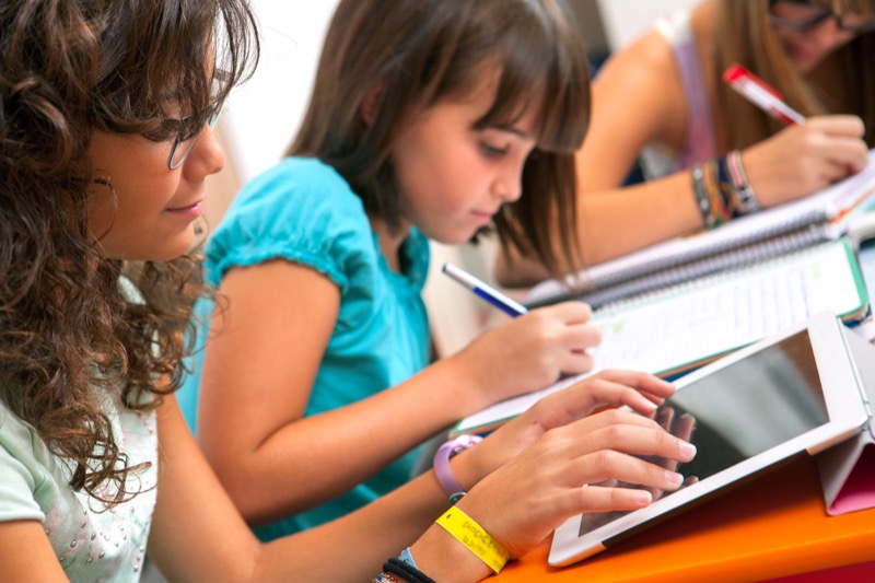 Epopeya La imagen muestra a 2 niñas, una escribiendo en su cuaderno y otra interactuando con la tablet