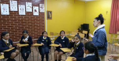 La imagen muestra un círculo de estudiantes con una profesora 