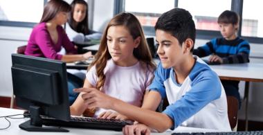 La imagen muestra a dos jóvenes leyendo o trabajando desde un computador una leyenda