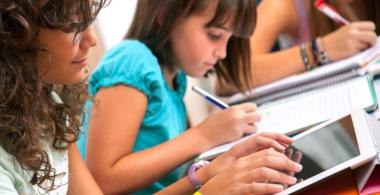 La imagen muestra a 2 niñas, una escribiendo en su cuaderno y otra interactuando con la tablet