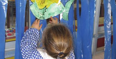 La imagen muestra a una niña pegando un dibujo de un pez en un fondo de plástico azul